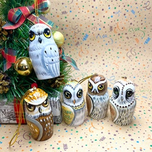 Peut inclure: Cinq décorations d'oiseaux en bois peintes à la main avec des motifs différents. Les hiboux sont tous blancs avec des accents bruns, noirs et jaunes. Ils sont suspendus à un sapin de Noël.