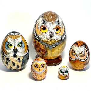 Peut inclure: Ensemble de cinq poupées russes en bois peintes à la main, chacune représentant un hibou avec des couleurs et des motifs différents. La plus grande poupée est brune, orange et noire avec un design détaillé. Les autres poupées sont plus petites et présentent différentes combinaisons de couleurs.