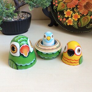 Peut inclure: Trois poup&eacute;es russes en bois peintes &agrave; la main en forme d'oiseaux. La plus grande poup&eacute;e est verte avec un visage blanc et un bec rouge. La poup&eacute;e du milieu est verte avec un oiseau bleu &agrave; l'int&eacute;rieur. La plus petite poup&eacute;e est jaune avec un bec noir et des accents orange.