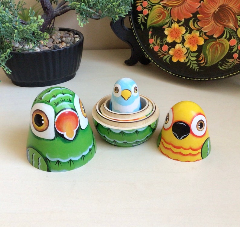 Peut inclure: Trois poup&eacute;es russes en bois peintes &agrave; la main en forme d'oiseaux. La plus grande poup&eacute;e est verte avec un visage blanc et un bec rouge. La poup&eacute;e du milieu est verte avec un oiseau bleu &agrave; l'int&eacute;rieur. La plus petite poup&eacute;e est jaune avec un bec noir et des accents orange.