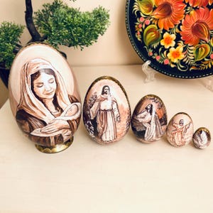 Peut inclure: Un ensemble de cinq œufs de Pâques en bois peints. Le plus grand œuf représente une femme tenant un bébé, tandis que les autres œufs présentent des figures religieuses. Les œufs sont peints dans des tons de marron, beige et or, avec des détails complexes.