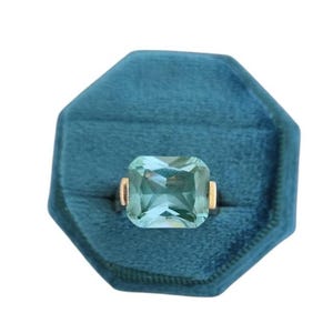 May include: A gold ring with a large, rectangular, light blue gemstone. The ring is set in a teal velvet ring box. The gemstone is faceted, reflecting light. The ring's band has gold accents on either side of the gemstone.