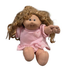 May include: A vintage Cabbage Patch Kid doll with blonde hair and a pink dress. The doll has a heart-shaped patch on the chest and a ruffled hem. The doll's arms and legs are made of a soft, flesh-toned material.
