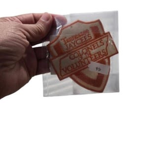 Könnte beinhalten: Ein schildförmiger, orange-weißer Stickaufnäher in einer transparenten Kunststoffhülle. Der Aufnäher trägt den Text Tennessee Jaycees und Colonels of the Volunteers. Ein kleiner weißer Aufkleber mit der handschriftlichen Zahl 75 ist angebracht.