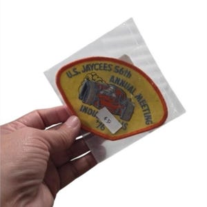 May include: A yellow embroidered patch with an orange border featuring a red race car graphic and the text U.S. JAYCEES 56th ANNUAL MEETING INDIANAPOLIS '76. The patch is enclosed in a clear plastic sleeve with a small white sticker labeled 45.