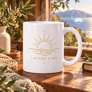 Könnte beinhalten: Ein weißer Keramikbecher mit einer goldfarbenen Grafik einer aufgehenden Sonne über Wellen. Unter der Grafik steht der Text I AM HERE & NOW. Das Design enthält vertikale gepunktete Linien oberhalb und unterhalb der Sonnen- und Wellendarstellung.