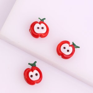 May include: Three red glass apple beads with white eyes and green stems. The beads are on a white surface.