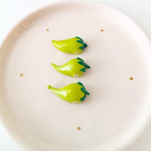 May include: Three small, green glass chilli pepper figurines with dark green stems and a glossy finish. The peppers are arranged on a light pink plate with three small gold dots.