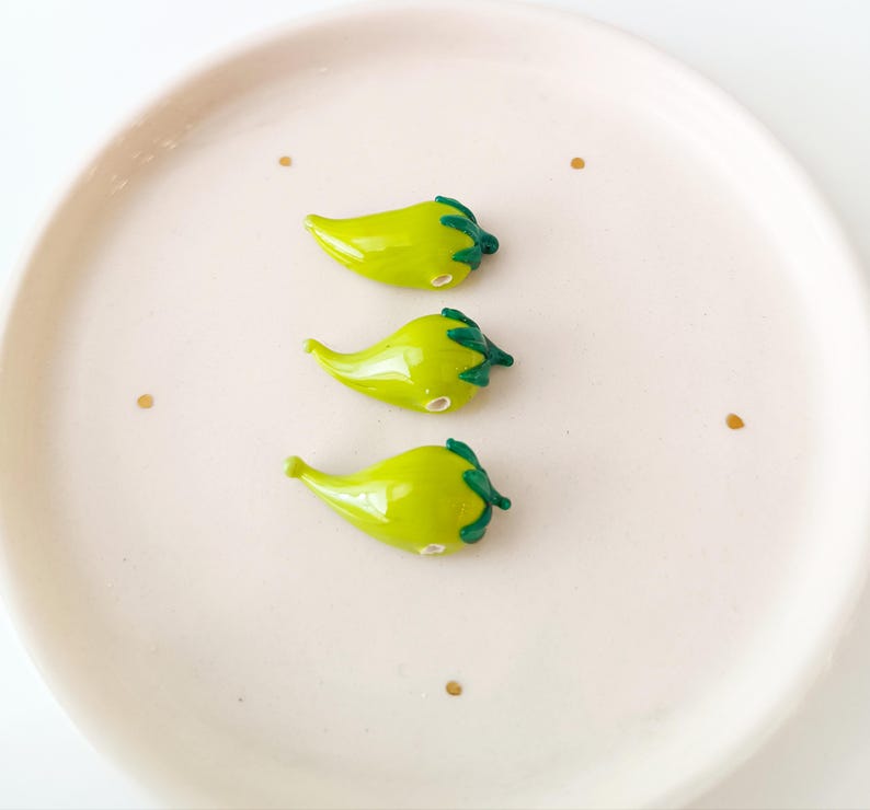 May include: Three small, green glass chilli pepper figurines with dark green stems and a glossy finish. The peppers are arranged on a light pink plate with three small gold dots.