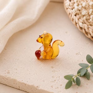Puede incluir: Una pequeña figura de ardilla hecha de vidrio translúcido de color ámbar. La ardilla está representada en posición sentada, sosteniendo una nuez de vidrio rojo oscuro entre sus patas delanteras. Tiene una cola curva, cabeza redondeada y ojos oscuros.