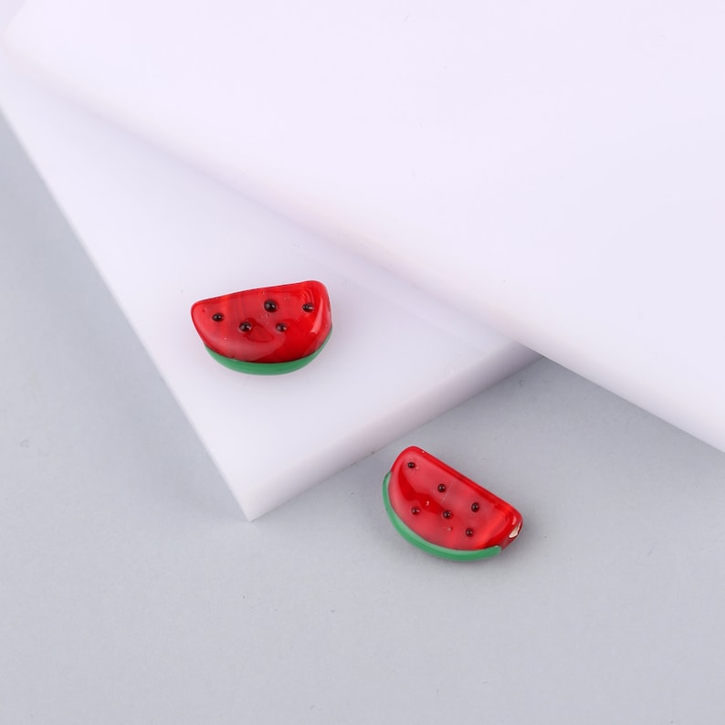 May include: Two small, wedge-shaped watermelon charms made of glossy red and green material. Each piece features a bright red top section with small, dark, circular spots representing seeds and a solid green base, mimicking the rind of a watermelon.