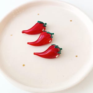 May include: Three glossy red chilli pepper ornaments with green stems and white accents rest on a cream-coloured plate with gold flecks. The peppers are arranged diagonally, showcasing their vibrant colour and detail.