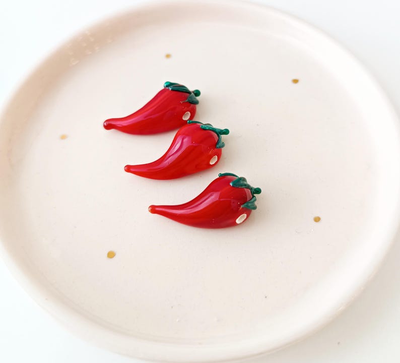 May include: Three glossy red chilli pepper ornaments with green stems and white accents rest on a cream-coloured plate with gold flecks. The peppers are arranged diagonally, showcasing their vibrant colour and detail.