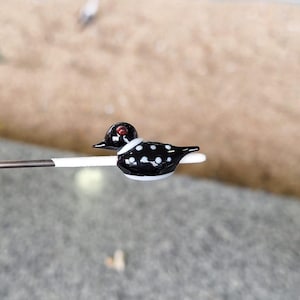 Puede incluir: Una figura de pato de cristal en miniatura. El pato tiene un cuerpo negro con lunares blancos, una cabeza negra y un ojo rojo. Tiene un collar blanco y est&aacute; posado sobre un palito blanco. El fondo est&aacute; borroso.
