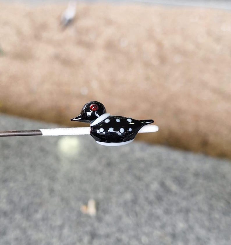 Puede incluir: Una figura de pato de cristal en miniatura. El pato tiene un cuerpo negro con lunares blancos, una cabeza negra y un ojo rojo. Tiene un collar blanco y est&aacute; posado sobre un palito blanco. El fondo est&aacute; borroso.