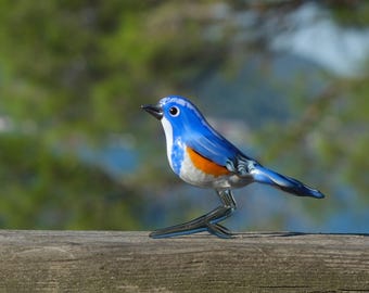 Può includere: Una statuetta di uccello in vetro blu e arancione con la pancia bianca. L'uccello è appollaiato su una superficie di legno.