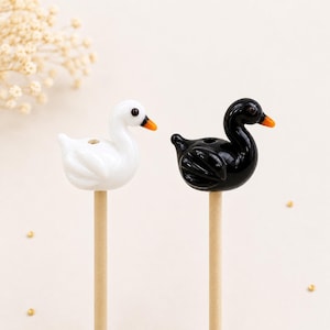 Puede incluir: Dos figuras decorativas de cisnes, una blanca y otra negra, cada una sobre un palito de madera. Los cisnes tienen picos naranjas y están sobre un fondo suave y neutro. El cisne blanco tiene un acabado brillante, mientras que el cisne negro tiene un aspecto liso y pulido.
