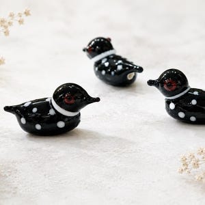 Puede incluir: Tres figuras decorativas de vidrio de somorgujo. Cada somorgujo negro tiene lunares blancos, un collar blanco y ojos rojos. Las figuras est&aacute;n dispuestas sobre una superficie blanca con delicados acentos florales.