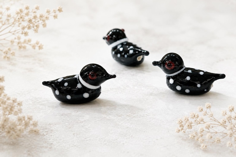 Puede incluir: Tres figuras decorativas de vidrio de somorgujo. Cada somorgujo negro tiene lunares blancos, un collar blanco y ojos rojos. Las figuras est&aacute;n dispuestas sobre una superficie blanca con delicados acentos florales.