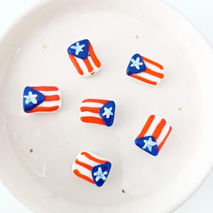 Könnte beinhalten: Sechs kleine Perlen mit dem Design der puerto-ricanischen Flagge. Jede Perle zeigt einen weißen Stern auf einem blauen Dreieck mit roten und weißen Streifen. Die Perlen liegen auf einer weißen, runden Oberfläche.