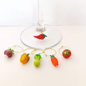 May include: A selection of wine glass charms featuring miniature vegetable designs. The charms include a red chilli pepper, tomato, carrot, and other vegetables, each suspended from a gold-coloured ring. These are designed to be attached to wine glasses.