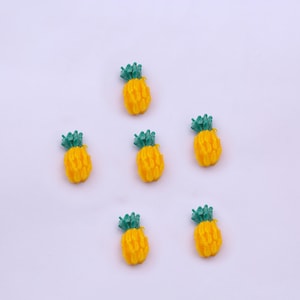 Könnte beinhalten: Sechs kleine gelbe Ananas-förmige Dekorationen mit grünen Spitzen. Die Ananas sind symmetrisch auf einem weißen Hintergrund angeordnet.