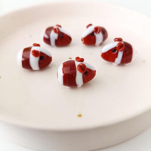 May include: Five miniature glass guinea pig figurines. Each figurine is red and white striped with small red ears and tiny black eyes. The figurines are arranged on a light pink plate.