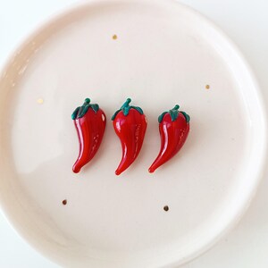 May include: Three glossy red chilli pepper-shaped decorative items with green stems and leaves are arranged on a white plate. The peppers have a smooth, curved shape and are positioned close together. The plate has a slightly raised edge.