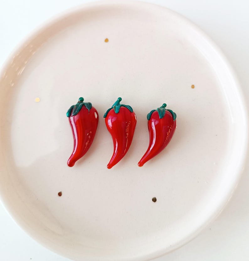 May include: Three glossy red chilli pepper-shaped decorative items with green stems and leaves are arranged on a white plate. The peppers have a smooth, curved shape and are positioned close together. The plate has a slightly raised edge.