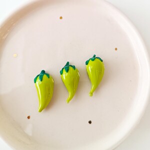 May include: Three small, glossy, lime-green chilli pepper-shaped objects with dark green stems are arranged on a light pink plate. The plate has a slightly raised edge and a few gold flecks scattered around the chillies.