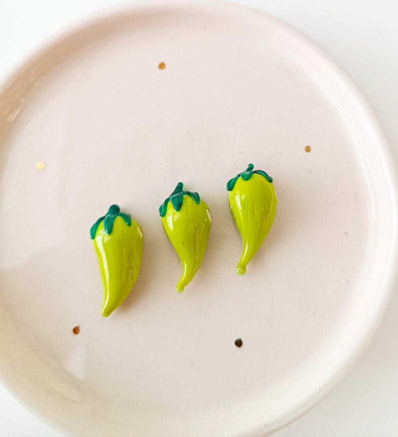 May include: Three small, glossy, lime-green chilli pepper-shaped objects with dark green stems are arranged on a light pink plate. The plate has a slightly raised edge and a few gold flecks scattered around the chillies.