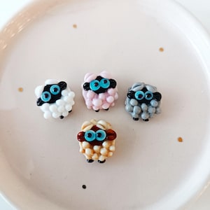 May include: Four colourful sheep figurines with blue eyes and black faces are arranged on a white plate. The sheep are white, pink, grey, and tan. The plate has small gold flecks.