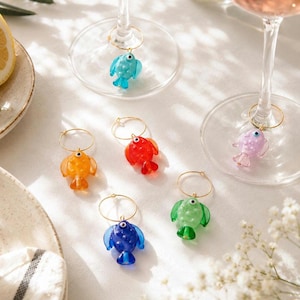 May include: A selection of colourful fish-shaped wine charms. Each charm is a different colour, including blue, orange, red, green, and purple. The charms are attached to gold-coloured hoops, designed to hang on a wine glass stem.