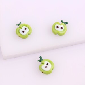 May include: Three green glass apple beads with white centers and brown eyes. The beads have a green stem on top.