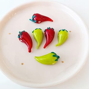 May include: A selection of red and green glass chili pepper ornaments, displayed on a white plate. The peppers have a shiny surface and detailed stems.