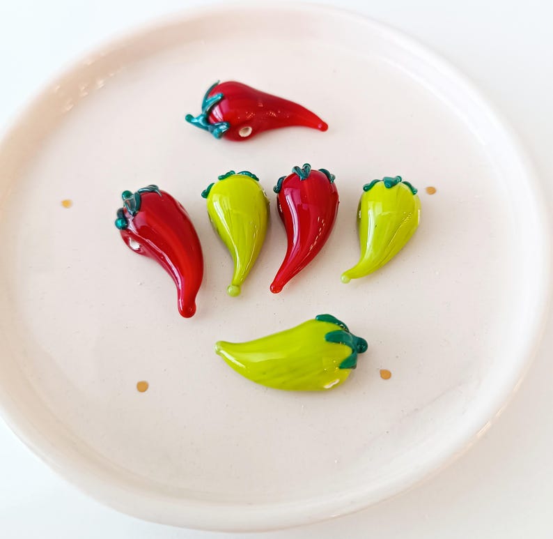 May include: A selection of red and green glass chili pepper ornaments, displayed on a white plate. The peppers have a shiny surface and detailed stems.