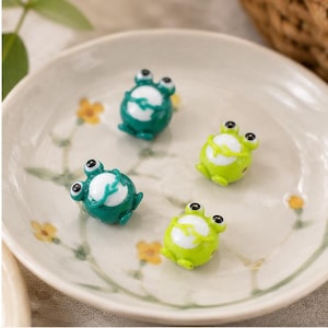Puede incluir: Cuatro pequeñas figuras con forma de rana en tonos verdes y amarillos están dispuestas sobre un plato decorativo. Cada rana tiene ojos grandes y negros y una barriga blanca. El plato tiene un diseño floral.