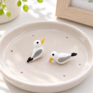 May include: Two small, white glass bird figurines with yellow beaks, grey wing details, and black tail tips. The birds are resting inside a shallow, round, off-white ceramic dish decorated with small, evenly spaced gold-coloured dots.