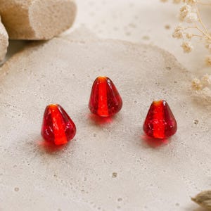 May include: Three small, teardrop-shaped glass beads in a vibrant, translucent red color. Each bead features a smooth, polished surface with a distinct vertical stripe of a slightly brighter, more intense red running through the center of the form.