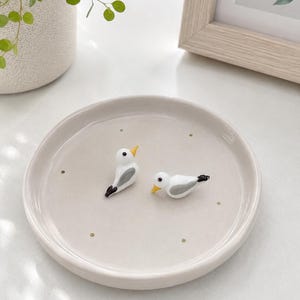 Op de afbeelding: Een ronde, lichtbeige keramische schaal met een gladde, glanzende afwerking en kleine, gelijkmatig verdeelde gouden stippen. Twee kleine glazen vogelbeeldjes in wit, grijs en zwart met gele snavels staan in het midden van de schaal.