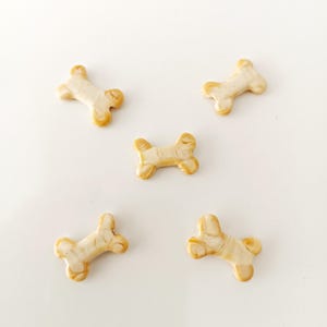 May include: Five small, bone-shaped decorative items. The bones are off-white with a marbled, tan-colored pattern and a golden-yellow trim. They are arranged on a white surface.