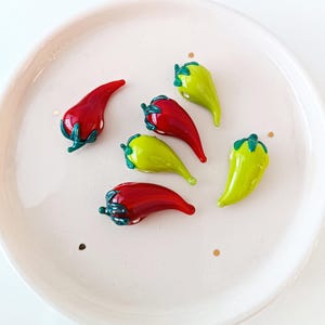 May include: A selection of glass chili pepper ornaments in red and yellow-green, with green stems, presented on a white plate. The peppers have a glossy finish and appear handmade.