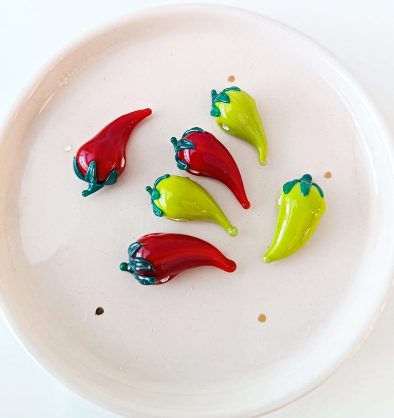 May include: A selection of glass chili pepper ornaments in red and yellow-green, with green stems, presented on a white plate. The peppers have a glossy finish and appear handmade.