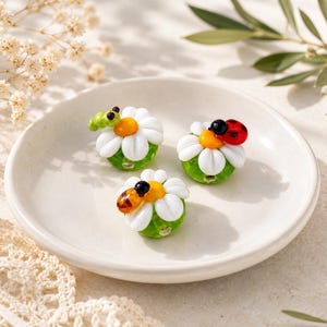 May include: Three glass beads shaped like white daisies with yellow centers, each featuring a small creature on top: a green caterpillar, a red ladybug with black spots, and a brown and black bee, all resting on a small white ceramic plate.