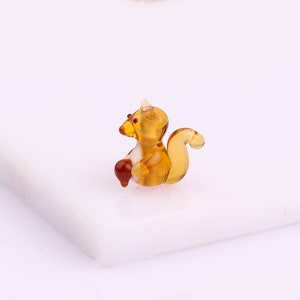 Peut inclure: Une petite figurine de  écureuil en verre de couleur brune et ambrée. L'écureuil tient un petit gland dans ses pattes.