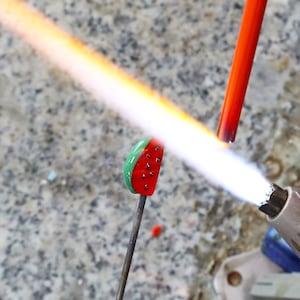 May include: A small glass watermelon slice charm being heated by a torch flame. The charm features a green rind, red flesh, and black seeds. A thin metal rod holds the glass piece while a bright orange flame from a torch melts the glass surface.