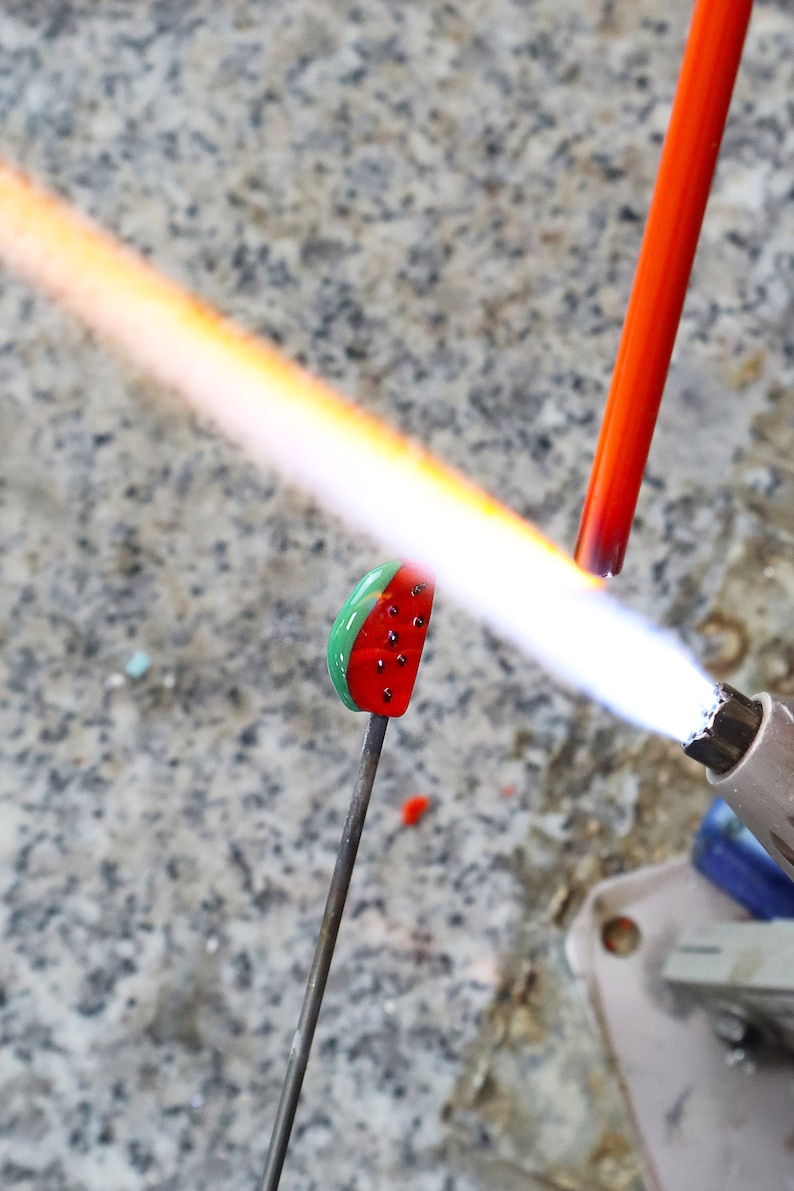 May include: A small glass watermelon slice charm being heated by a torch flame. The charm features a green rind, red flesh, and black seeds. A thin metal rod holds the glass piece while a bright orange flame from a torch melts the glass surface.