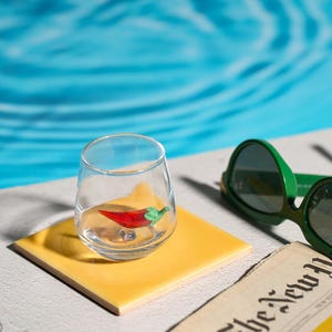 May include: A clear glass with a red and green chili pepper figurine inside, resting on a yellow square tile. Green sunglasses and a newspaper are in the background, with a blue water backdrop.