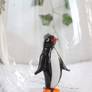 May include: A small, glass penguin figurine with a black body, white chest, red beak, and orange feet. The penguin is inside a clear glass container, creating a whimsical decorative piece.