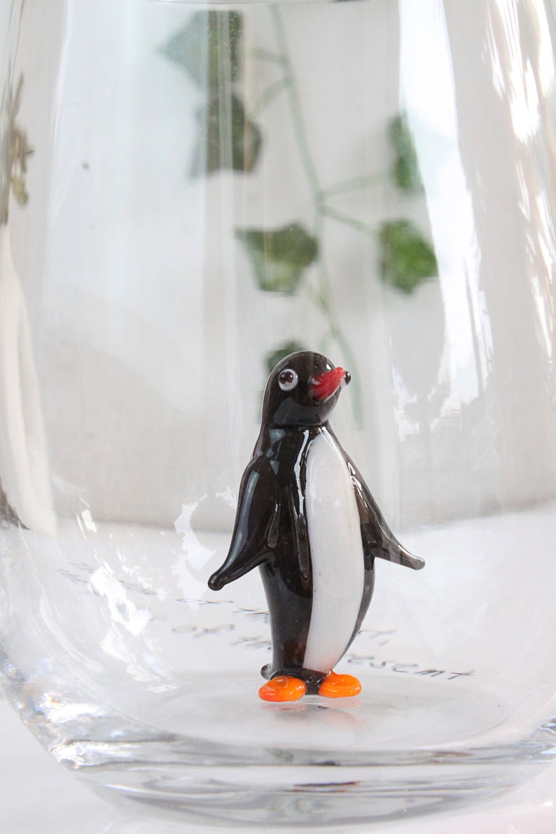 May include: A small, glass penguin figurine with a black body, white chest, red beak, and orange feet. The penguin is inside a clear glass container, creating a whimsical decorative piece.